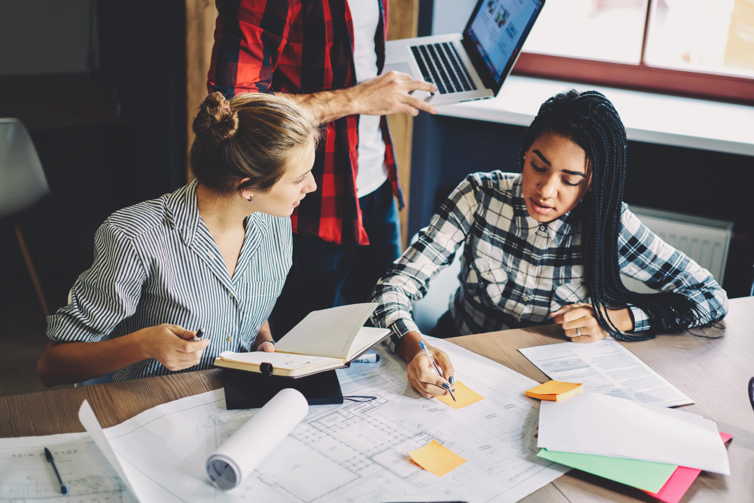 Skilled crew of male and female architect working on new project drawing blueprint of future building making last preparation before creating web model on application installed on laptop computer