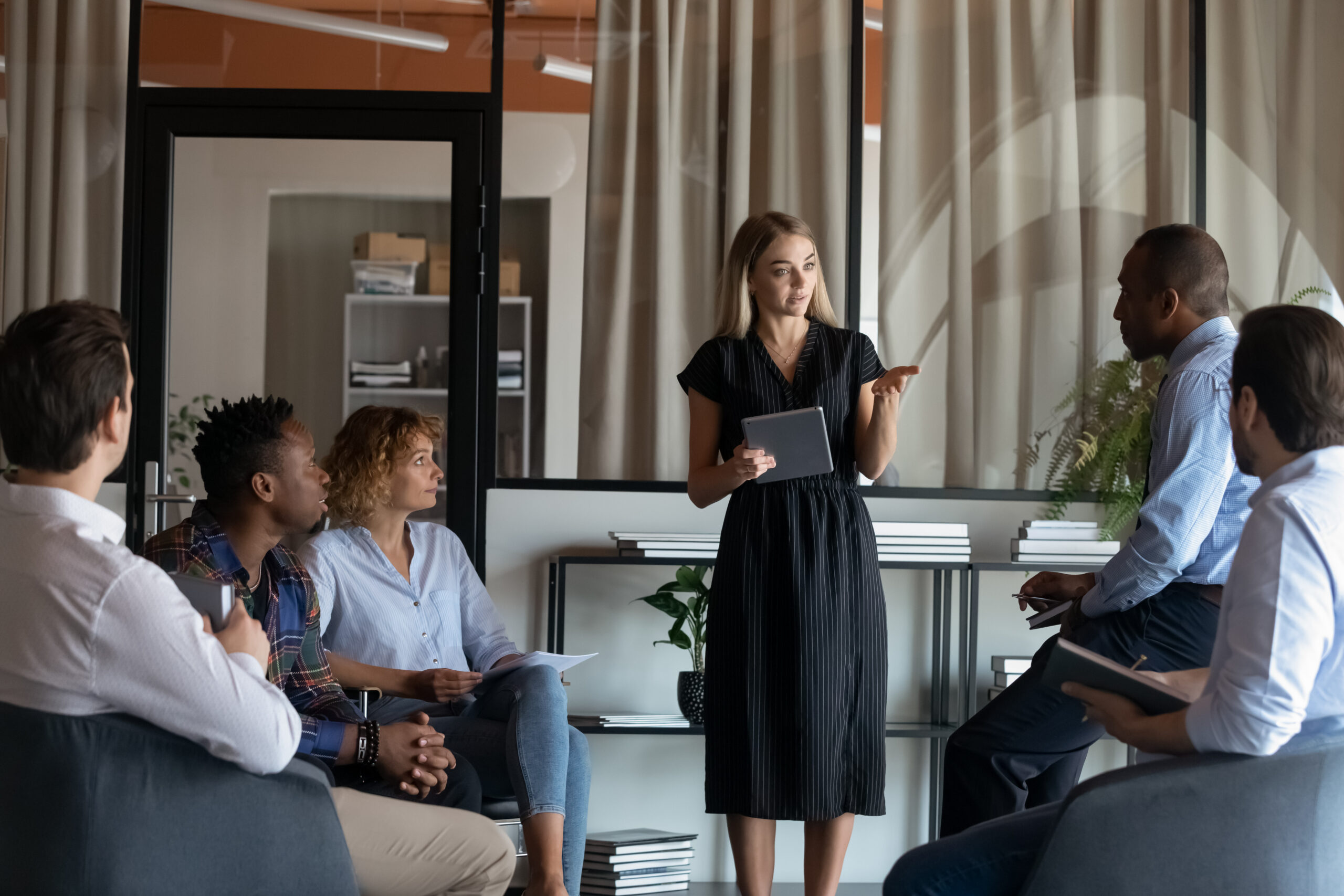 Confident businesswoman mentor holding tablet leading corporate meeting briefing with diverse employees in modern office boardroom, colleagues coworkers listening to executive, sitting in circle